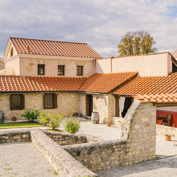 Steinhof mit Topfpflanzen und Fässern, umgeben von Gebäuden mit roten Ziegeldächern unter einem teilweise bewölkten Himmel.