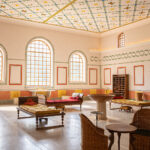 Ein geräumiger Raum mit gewölbten Fenstern, dekorativer Decke und Wänden, Liegestühlen, Korbstühlen, einem zentralen Steinbecken und Holzregalen an einer Wand.