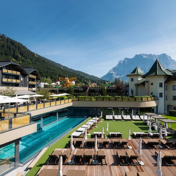 Luxushotel mit Außenpool, Liegestühlen und Terrasse inmitten von bewaldeten Hügeln und einem Berg unter blauem Himmel.
