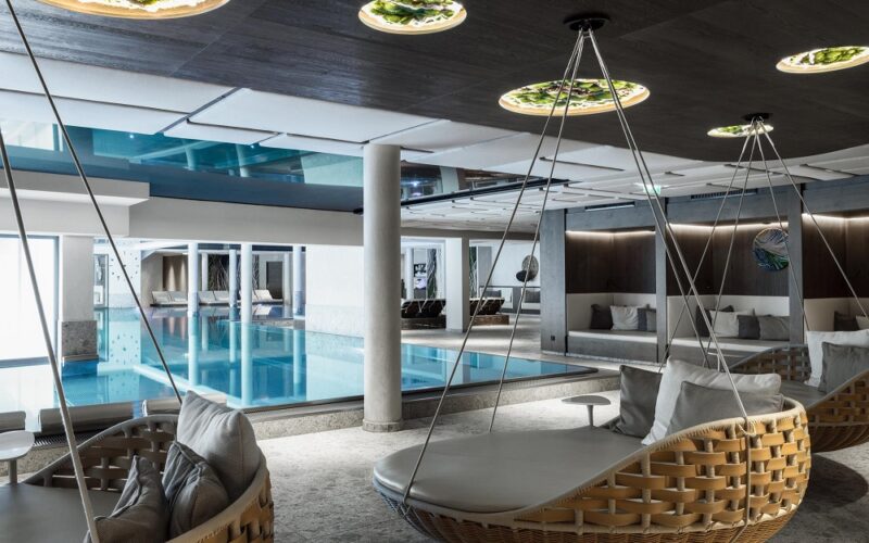 Moderner Hallenbadbereich mit großen gepolsterten Schaukelstühlen, Glaswänden, einem Becken mit blauem Wasser und Deckenleuchten, die einen eleganten Freizeitbereich schaffen.
