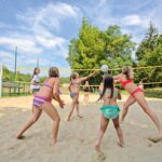 Fünf Mädchen in Badeanzügen spielen Volleyball auf einem sandigen Platz im Freien, mit Bäumen und blauem Himmel im Hintergrund.