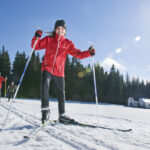 Ein Kind in einer roten Jacke fährt Langlaufski im Schnee, gefolgt von drei Personen, unter einem hellen, sonnigen Himmel in der Nähe eines Waldes und eines kleinen Gebäudes.