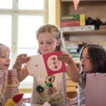 Eine Gruppe kleiner Kinder sitzt um einen Tisch herum, während ein Mädchen in der Mitte eine Grußkarte öffnet und vorliest.