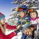 Eine Frau und drei Kinder in Winterkleidung lachen und spielen zusammen im Schnee mit einem verschneiten Baum im Hintergrund.