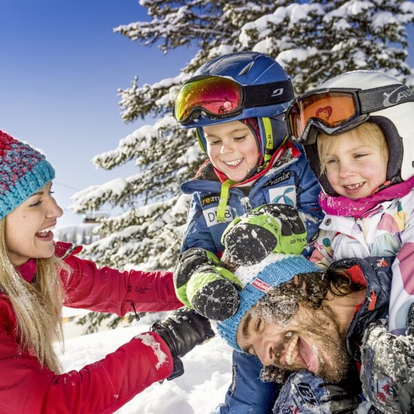 Eine Frau und drei Kinder in Winterkleidung lachen und spielen zusammen im Schnee mit einem verschneiten Baum im Hintergrund.