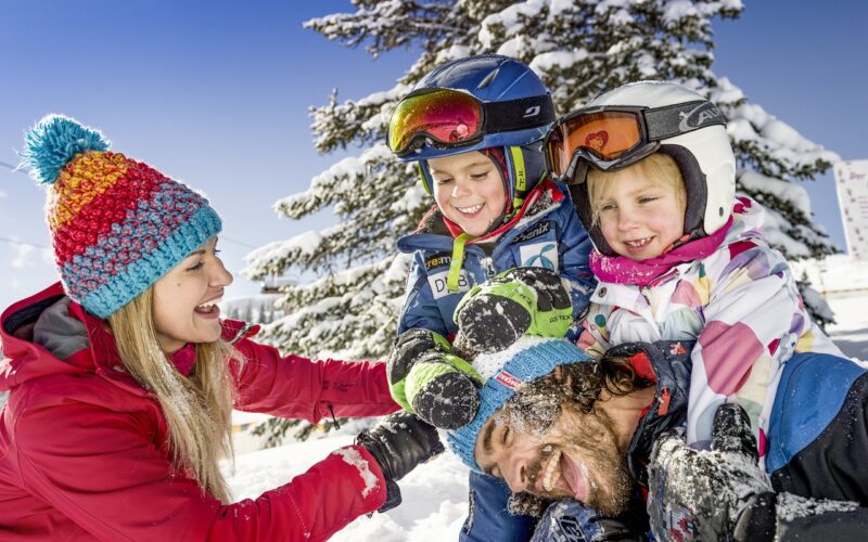 Eine Frau und drei Kinder in Winterkleidung lachen und spielen zusammen im Schnee mit einem verschneiten Baum im Hintergrund.