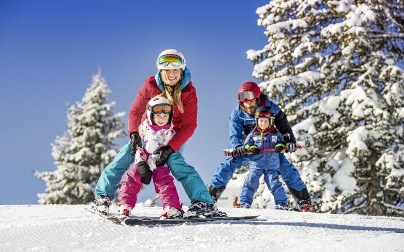 Eine vierköpfige Familie in Winterkleidung fährt gemeinsam auf einer verschneiten Piste mit schneebedeckten Bäumen und einem klaren blauen Himmel im Hintergrund Ski.
