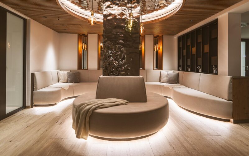 Moderner Lounge-Bereich mit geschwungenen beigen Sofas, einem runden, mit einer Decke drapierten Ottomanen in der Mitte, sanfter Raumbeleuchtung und Holzakzenten an Decke und Boden.