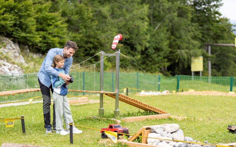 Ein Erwachsener hilft einem Kind, ein ferngesteuertes Auto auf einem von Gras und Bäumen umgebenen Hindernisparcours zu steuern.