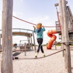 Ein Kind balanciert auf einer Seilbrücke auf einem Spielplatz im Freien, umgeben von Holzstrukturen und einer roten Spiralrutsche.