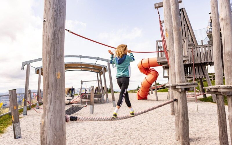 Ein Kind balanciert auf einer Seilbrücke auf einem Spielplatz im Freien, umgeben von Holzstrukturen und einer roten Spiralrutsche.