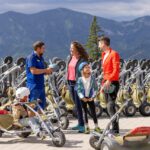 Eine Familie und ein Ausbilder stehen zwischen mehreren Mountain Trikes, halten Helme und unterhalten sich, während im Hintergrund die Berge zu sehen sind.