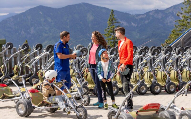 Eine Familie und ein Ausbilder stehen zwischen mehreren Mountain Trikes, halten Helme und unterhalten sich, während im Hintergrund die Berge zu sehen sind.