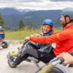 Zwei Personen mit Helmen fahren mit einem Go-Kart auf einem Schotterweg mit Bergen und Bäumen im Hintergrund. Ein weiterer Fahrer ist in der Ferne zu sehen.