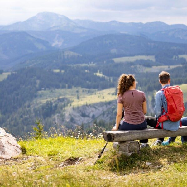 Drei Personen sitzen auf einer Holzbank mit Blick auf eine Berglandschaft, neben ihnen eine Wanderausrüstung und in der Nähe auf einem Felsen ein Wegweiser.