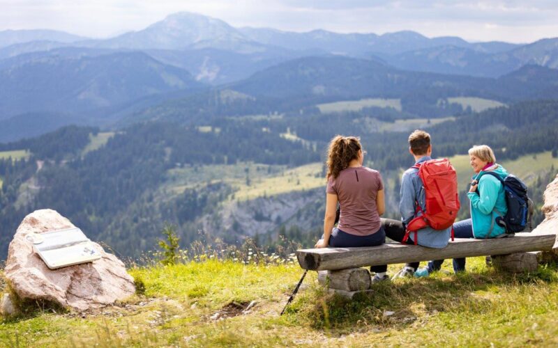 Drei Personen sitzen auf einer Holzbank mit Blick auf eine Berglandschaft, neben ihnen eine Wanderausrüstung und in der Nähe auf einem Felsen ein Wegweiser.