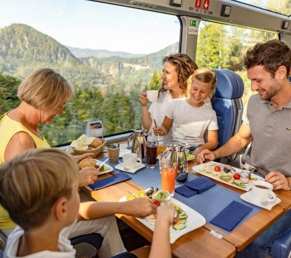 Eine fünfköpfige Familie genießt eine gemeinsame Mahlzeit an einem Tisch in einem modernen Zug mit großen Fenstern und Blick auf eine malerische Berglandschaft.