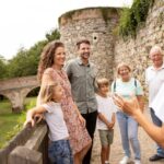 Eine Gruppe von sechs Personen, darunter zwei Kinder, steht lächelnd in der Nähe einer Steinmauer und einer Brücke, während eine Frau im Vordergrund im Freien zu ihnen spricht.