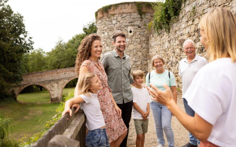 Eine Gruppe von sechs Personen, darunter zwei Kinder, steht lächelnd in der Nähe einer Steinmauer und einer Brücke, während eine Frau im Vordergrund im Freien zu ihnen spricht.