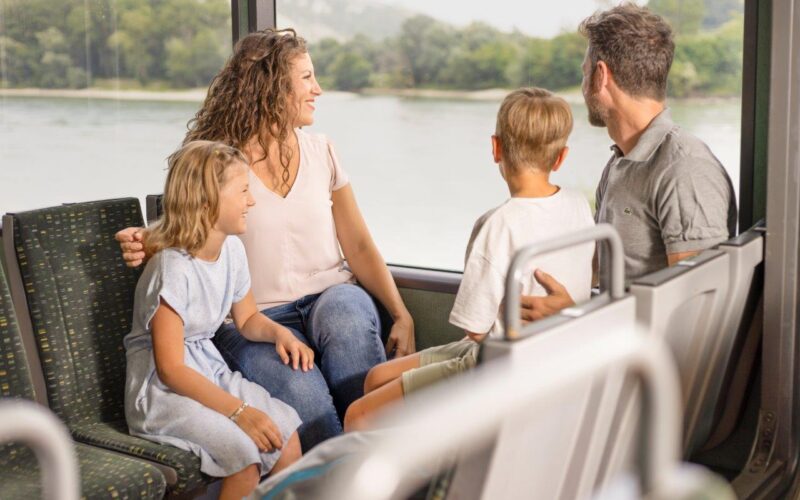 Eine vierköpfige Familie sitzt gemeinsam in einem Bus und blickt aus dem Fenster auf eine malerische Landschaft mit Wasser und Grünflächen.