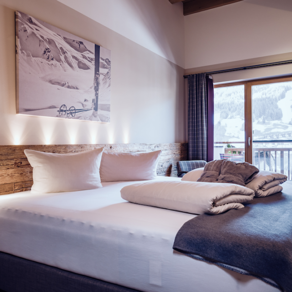 Modernes Hotelzimmer mit einem großen Bett, ordentlich gefalteten Handtüchern, einer Akzentwand aus Holz und einem Fenster mit Blick auf die verschneiten Berge draußen.