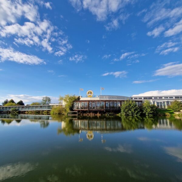 Ein Gebäude mit einer goldenen Löwenstatue auf dem Dach spiegelt sich in einem ruhigen See, umgeben von Bäumen und unter einem blauen Himmel mit vereinzelten Wolken.