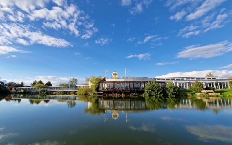 Ein Gebäude mit einer goldenen Löwenstatue auf dem Dach spiegelt sich in einem ruhigen See, umgeben von Bäumen und unter einem blauen Himmel mit vereinzelten Wolken.