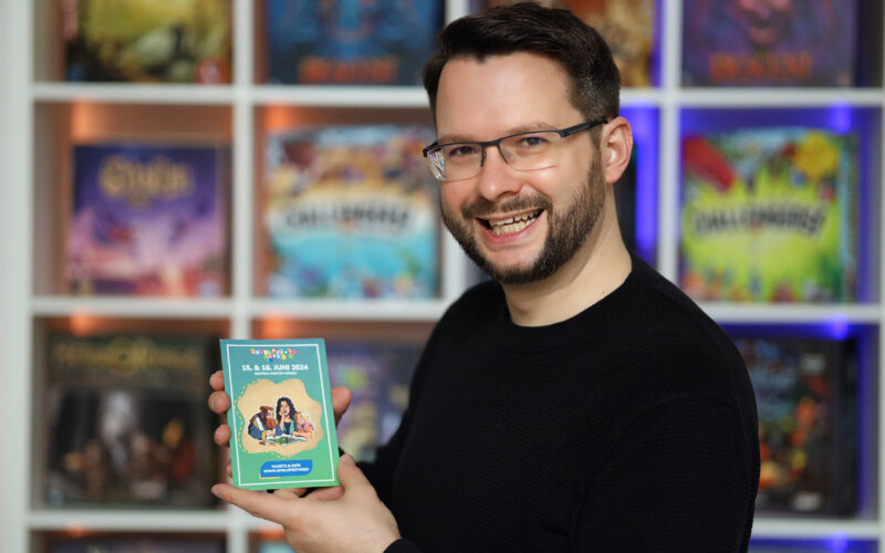 Ein Mann mit Brille und Bart lächelt in die Kamera und hält vor einem Regal voller Brettspiele ein buntes Veranstaltungsheft hoch.