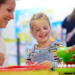 Ein junges Mädchen mit Gesichtsbemalung lächelt, während es mit einer erwachsenen Frau an einem Tisch im Haus ein Brettspiel spielt.