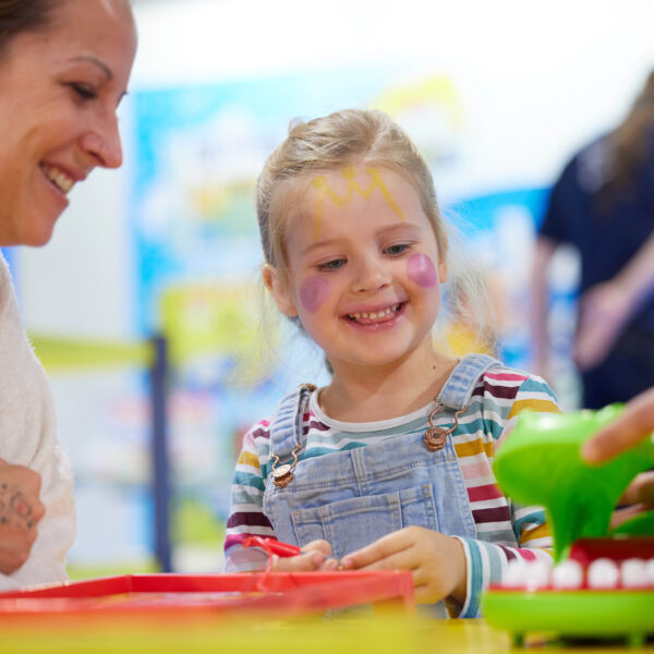 Ein junges Mädchen mit Gesichtsbemalung lächelt, während es mit einer erwachsenen Frau an einem Tisch im Haus ein Brettspiel spielt.