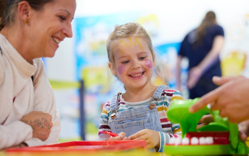 Ein junges Mädchen mit Gesichtsbemalung lächelt, während es mit einer erwachsenen Frau an einem Tisch im Haus ein Brettspiel spielt.