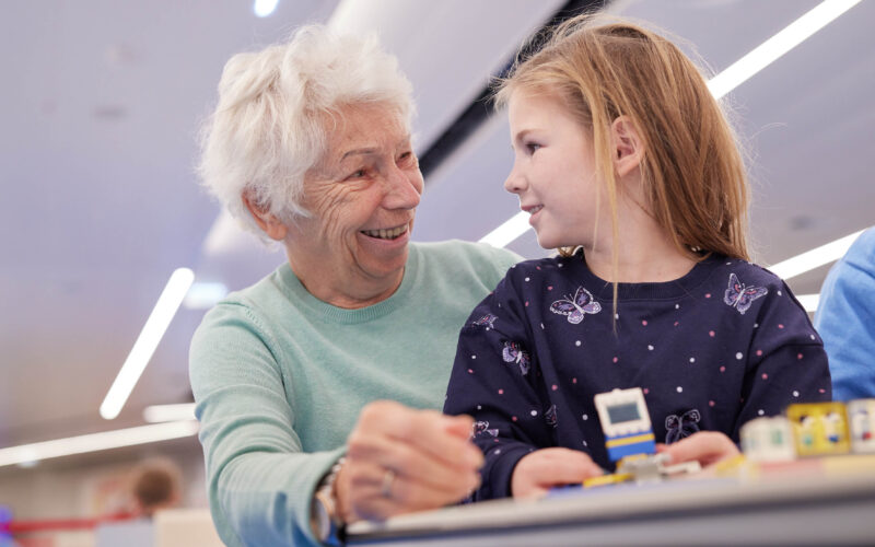 Eine ältere Frau und ein junges Mädchen sitzen zusammen im Haus, lächeln sich an, während sie an einem Tisch an einem kleinen Bauspielzeug arbeiten und ihr eigenes kleines Spielefest genießen.