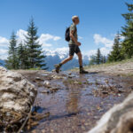 Eine Person mit einem Rucksack wandert auf einem felsigen Bergpfad in der Nähe eines seichten Baches, mit Bäumen und schneebedeckten Gipfeln im Hintergrund.