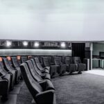 Ein moderner, runder Raum mit abgestuften Sitzgelegenheiten vor einer großen weißen, kuppelförmigen Leinwand, mit schwarzen und grauen Stühlen, einige mit orangefarbenen Akzenten.