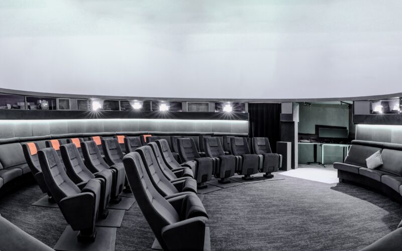 Ein moderner, runder Raum mit abgestuften Sitzgelegenheiten vor einer großen weißen, kuppelförmigen Leinwand, mit schwarzen und grauen Stühlen, einige mit orangefarbenen Akzenten.