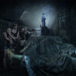 Eine Gruppe von Menschen geht durch einen dunklen, unheimlichen Raum mit bedeckten Körpern auf dem Boden, während eine gewandete Gestalt auf einem Podest am anderen Ende des Raumes steht und leuchtet.