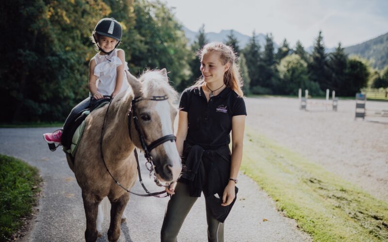 Ein kleines Kind mit einem Helm sitzt auf einem hellbraunen Pferd, während ein älteres Mädchen das Pferd auf einem Reitplatz im Freien führt.