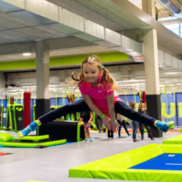 Ein junges Mädchen in Sportkleidung macht einen Spagatsprung über ein Trampolin in einem Indoor-Trampolinpark, im Hintergrund sind andere Leute zu sehen.