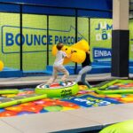 Drei Kinder spielen mit großen, gelben, gepolsterten Stöcken auf einer aufblasbaren Plattform in einem Indoor-Trampolin- und Aktivitätspark mit der Beschilderung „Bounce“ und „Jump Dome“.