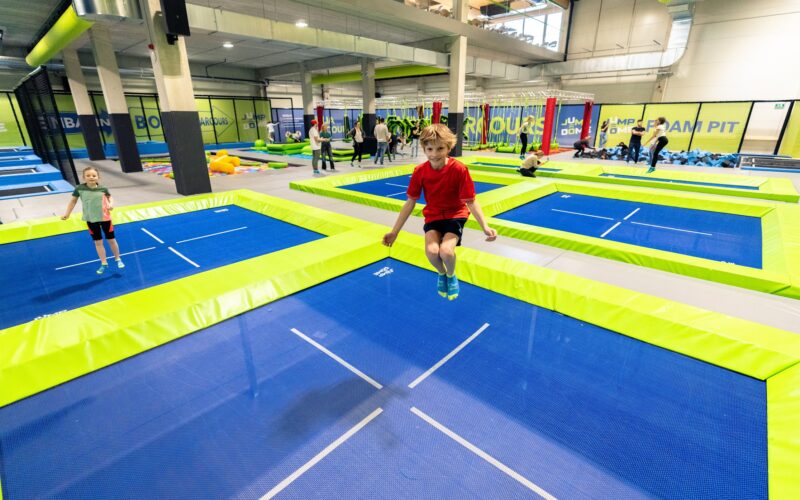 Kinder springen auf blauen und grünen Trampolinen in einem Indoor-Trampolinpark, im Hintergrund sind andere Personen und Geräte zu sehen.