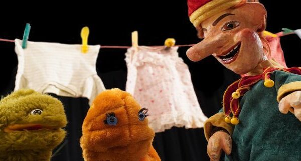 Vor einem dunklen Hintergrund stehen drei bunte Puppen vor einer Wäscheleine, an der Kinderkleidung hängt.