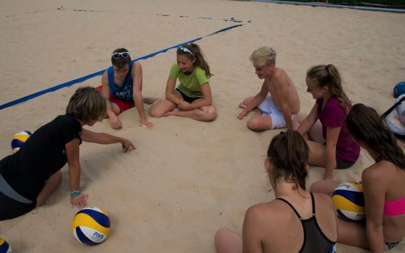 Eine Gruppe junger Leute sitzt im Kreis auf einem Beachvolleyballfeld und hört einem Trainer zu, der mit dem Finger in den Sand zeichnet. Mehrere Volleybälle liegen auf dem Boden.