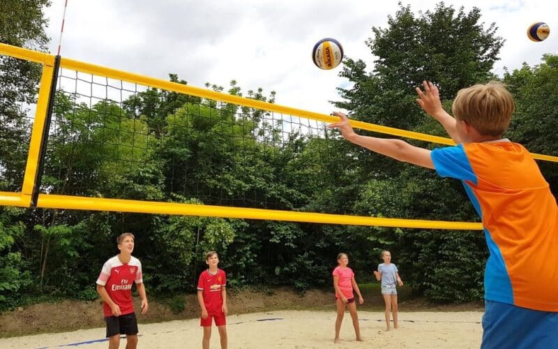 Fünf Kinder spielen Volleyball auf einem Sandplatz im Freien. Ein Spieler in einem orangefarbenen Trikot springt, um den Ball über das Netz zu schlagen. Im Hintergrund sind Bäume und ein bewölkter Himmel zu sehen.