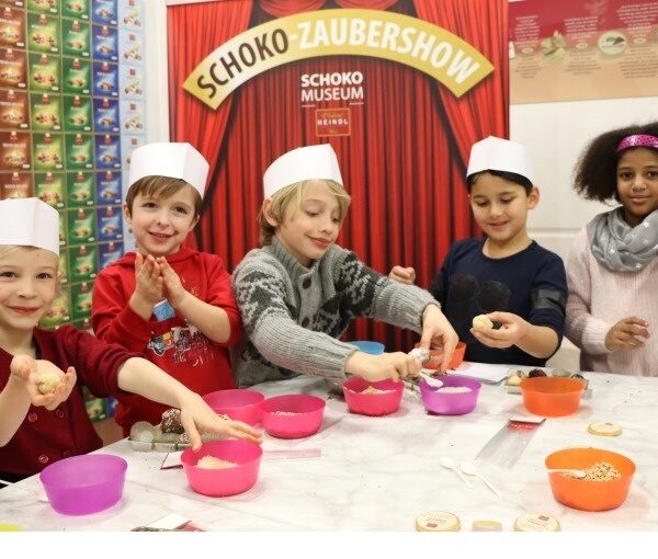Fünf Kinder mit Papierhüten stellen an einem Tisch im Schoko-Museum Schokoladenleckereien her. Im Hintergrund stehen Schüsseln mit Zutaten und eine Schautafel mit der Aufschrift „Schoko-Zaubershow“.
