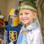 Kinder in mittelalterlichen Kostümen, mit einem lächelnden Mädchen im Vordergrund und einem Jungen in einem Ritterkostüm hinter ihr.