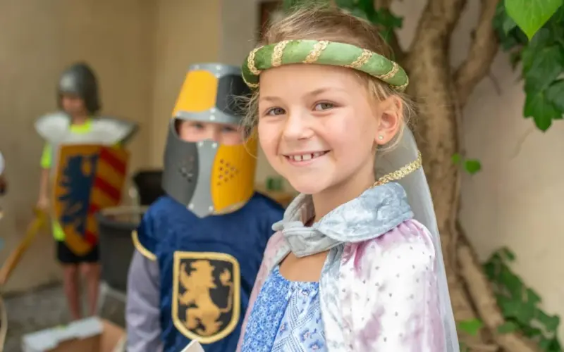 Kinder in mittelalterlichen Kostümen, mit einem lächelnden Mädchen im Vordergrund und einem Jungen in einem Ritterkostüm hinter ihr.