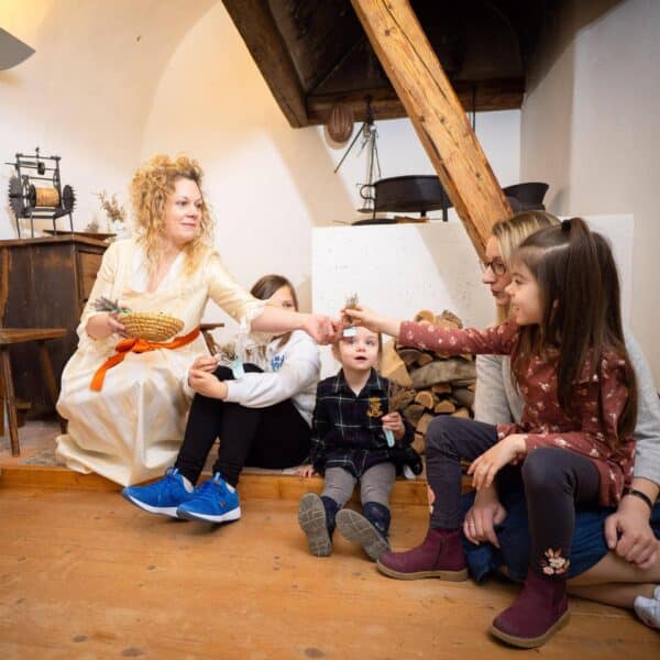 Eine Frau in einem weißen Kleid überreicht einem Mädchen, das mit anderen Kindern und einem Erwachsenen in einem rustikalen Raum auf dem Boden sitzt, einen kleinen Gegenstand.