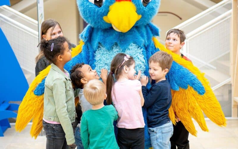 In einem Gebäude steht eine Gruppe Kinder herum und umarmt eine Person in einem großen, bunten blau-gelben Vogelkostüm.