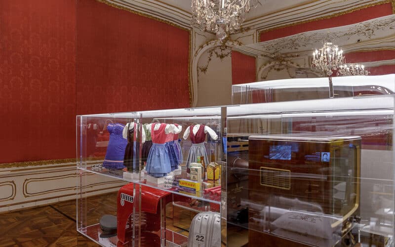 In einer Glasvitrine in einem reich verzierten Raum werden unter Kronleuchtern und roter Tapete alte Puppenkleider, Spielzeuge und Haushaltsgegenstände im Miniaturformat ausgestellt.