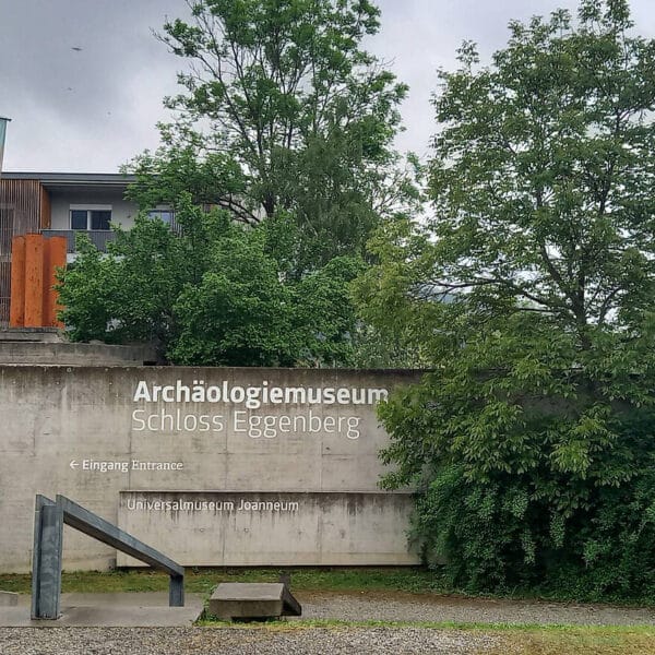 Betonwand mit Beschilderung „Archäologiemuseum Schloss Eggenberg“, Eingangsschild, Bannern, Bäumen und im Hintergrund ist ein Teil eines modernen Gebäudes zu sehen.